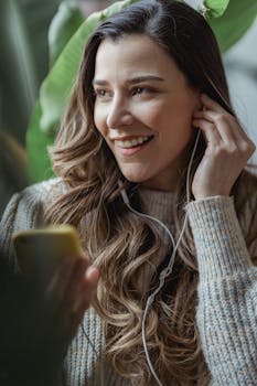 Cheerful young woman listening to music on her smartphone indoors, wearing warm sweater.