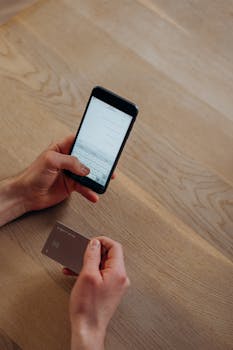A person making a cashless transaction using a smartphone and credit card on a wooden table.